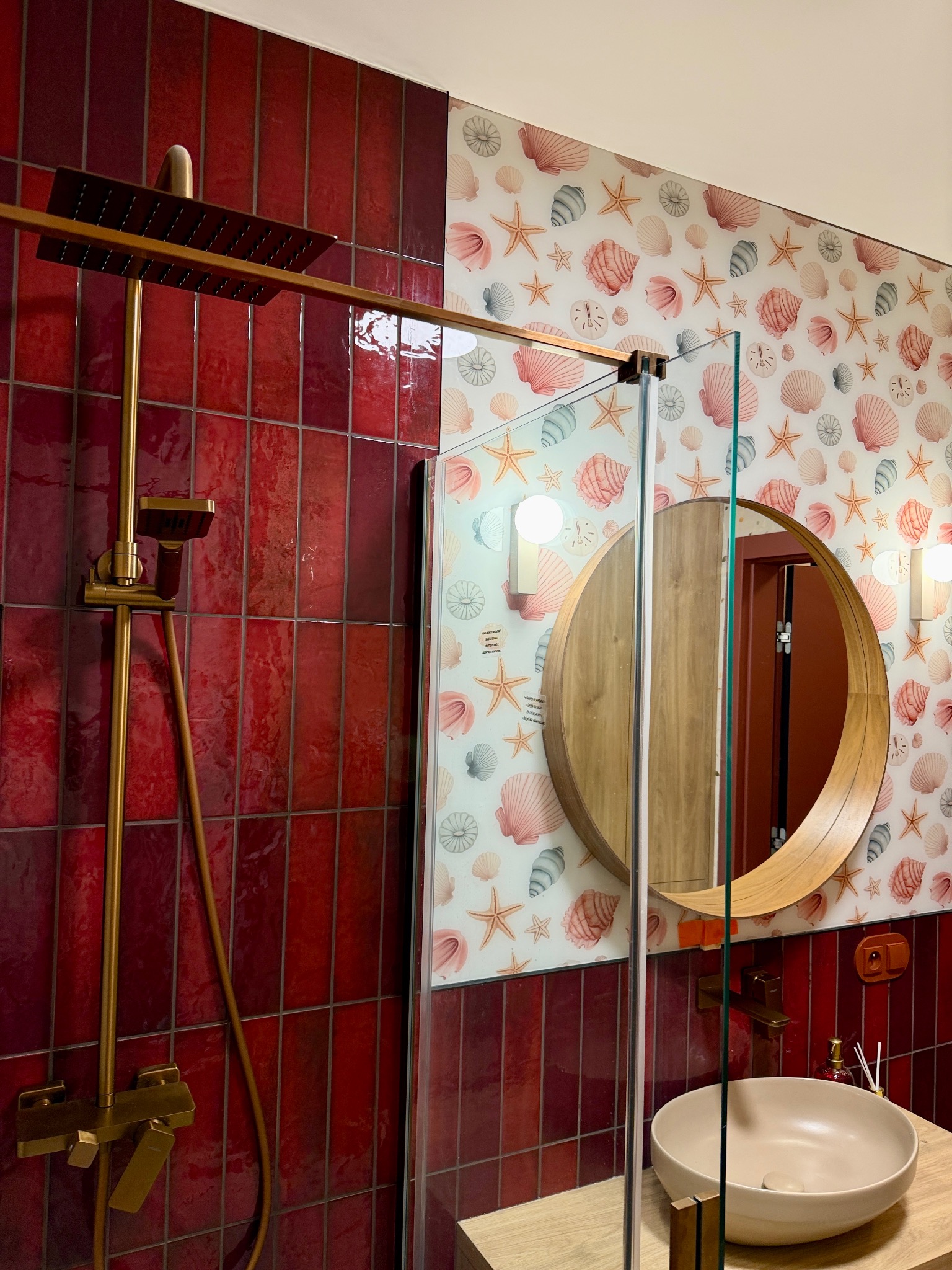 Nowoczesna łazienka z czerwonymi płytkami, złotą armaturą i okrągłym lustrem na ścianie w muszelki. Kabina prysznicowa ze szklanymi drzwiami.