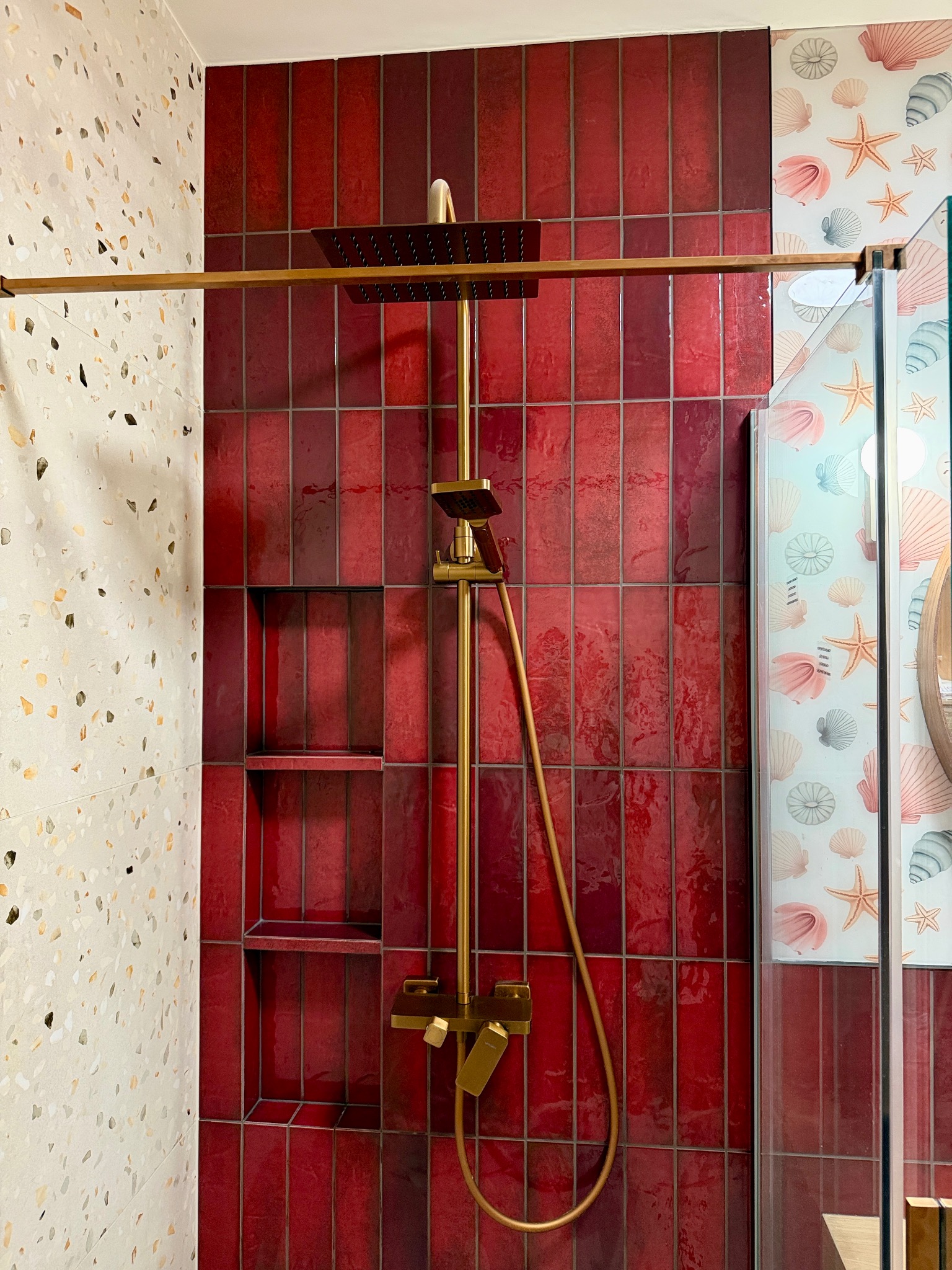 Nowoczesna kabina prysznicowa z czerwoną, błyszczącą glazurą ułożoną pionowo, złotą armaturą i półkami wnękowymi. Ściana obok wykończona płytkami lastryko.