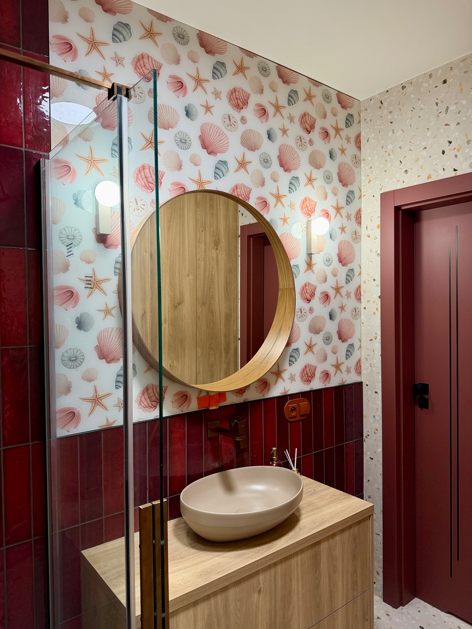 Wykończona łazienka z kabiną prysznicową, okrągłym lustrem i umywalką nablatową. Czerwone płytki i tapeta w muszelki dodają unikalnego charakteru.