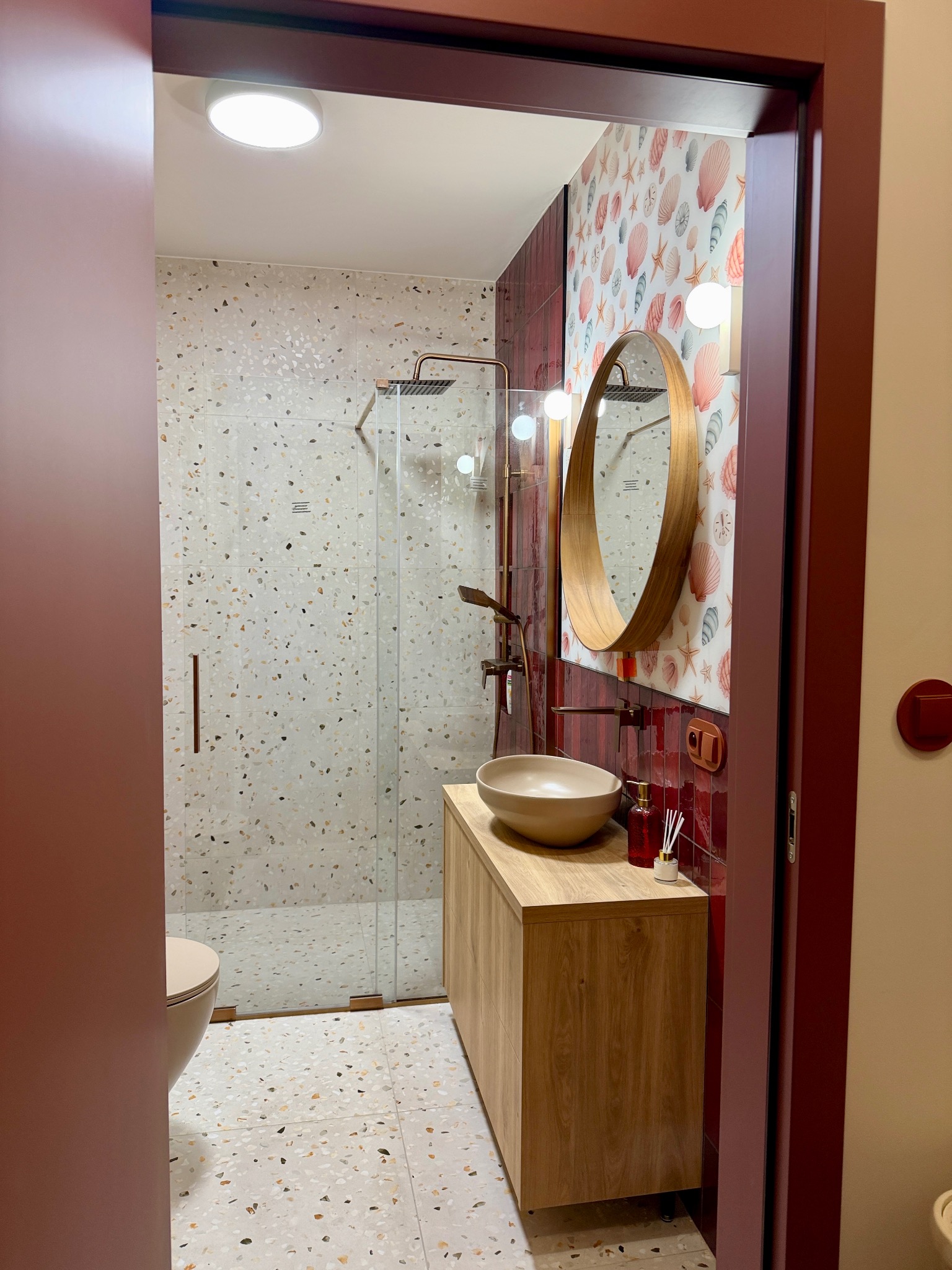 Nowoczesna łazienka z kabiną prysznicową, umywalką na drewnianej szafce i okrągłym lustrem. Ściany zdobią płytki w stylu lastryko i motywy morskie.