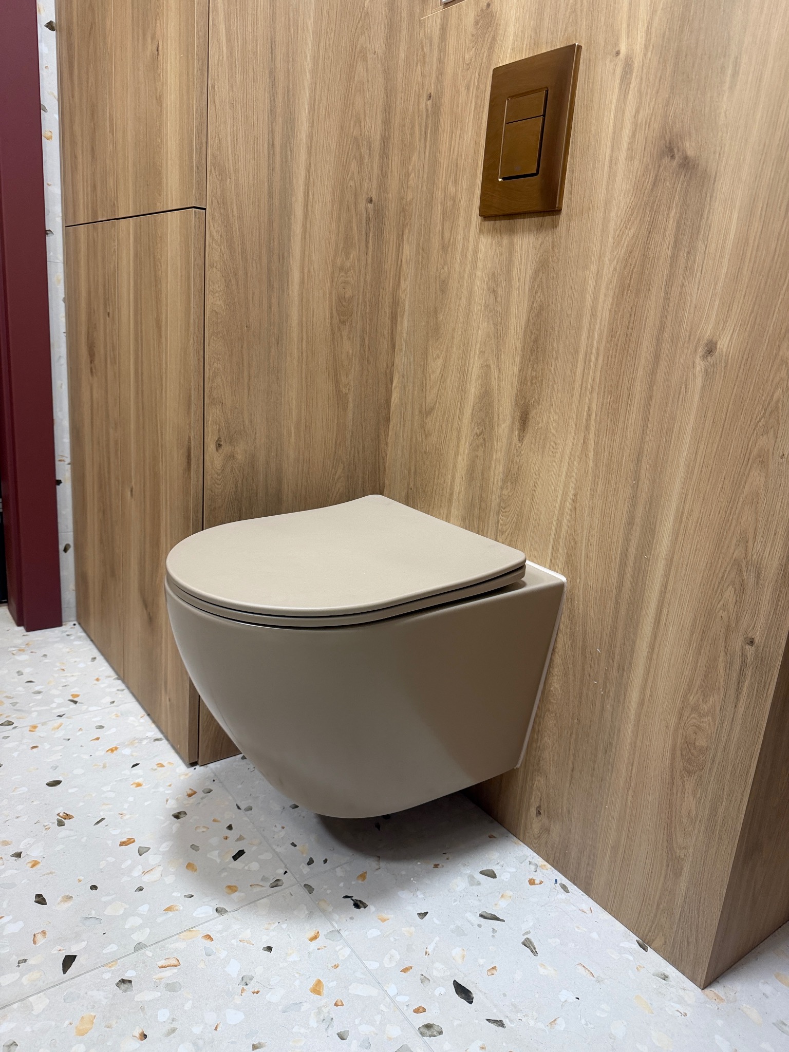 Beżowa, podwieszana toaleta w nowoczesnej łazience z drewnianą zabudową i mozaikową podłogą. Uwagę zwraca nietypowy, brązowy włącznik światła.