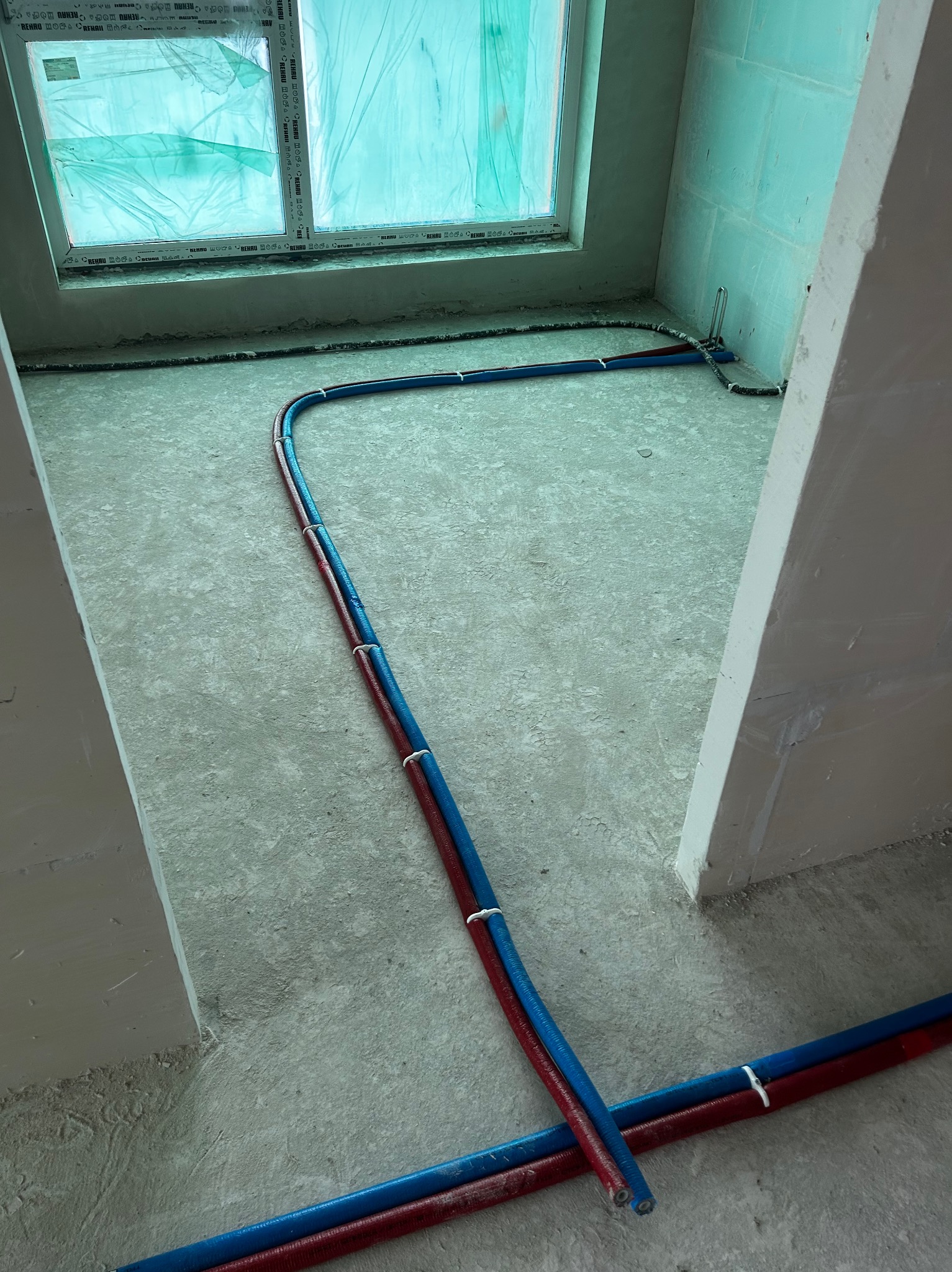 Instalacja rur (czerwona i niebieska) na podłodze w surowym wnętrzu, widok z góry. Rury przymocowane białymi opaskami. Okno z folią ochronną w tle.