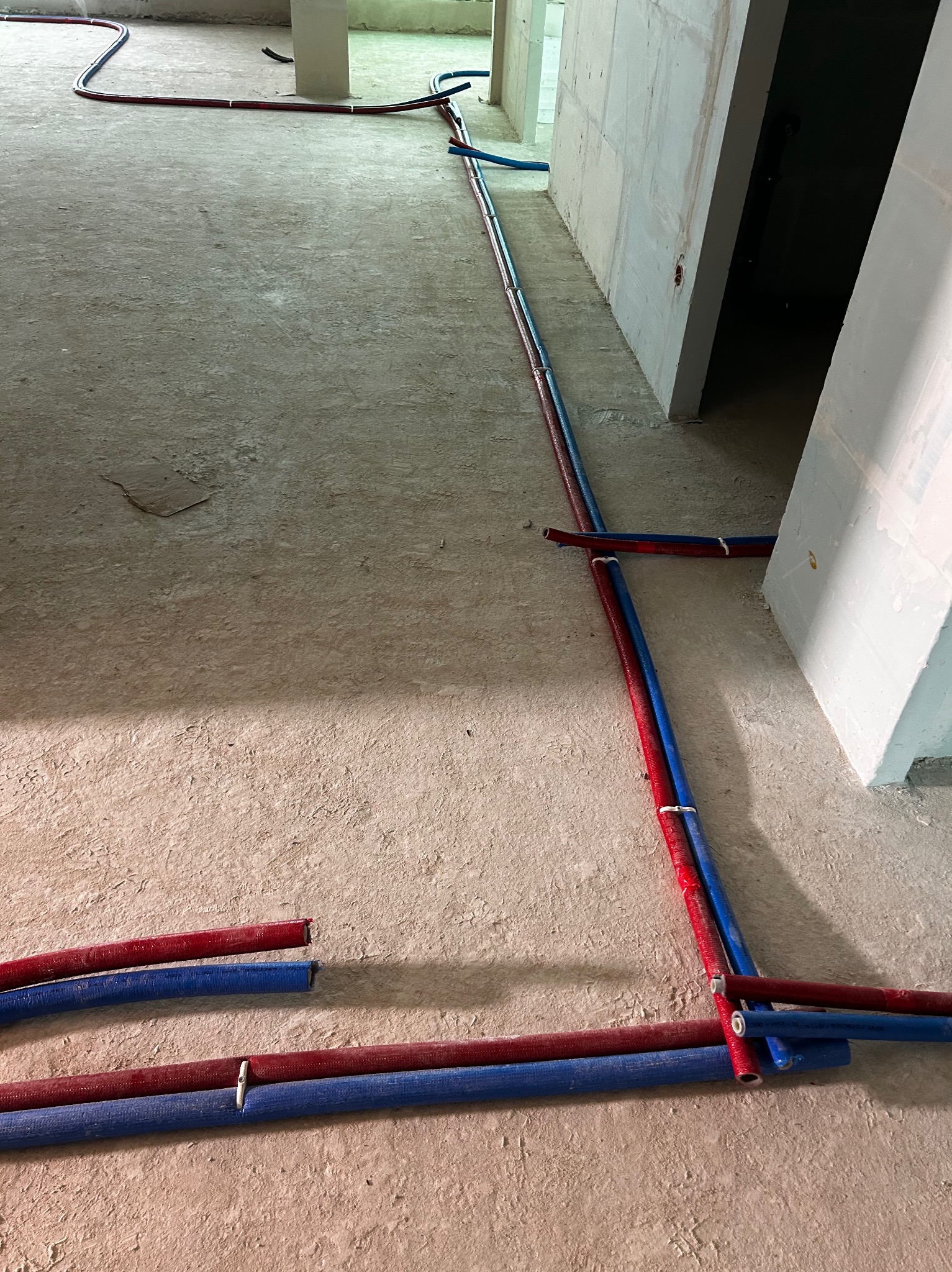 Niebiesko-czerwone rury instalacji grzewczej ułożone na surowym betonie w nowym budynku, widoczne połączenia i fragmenty ścian.