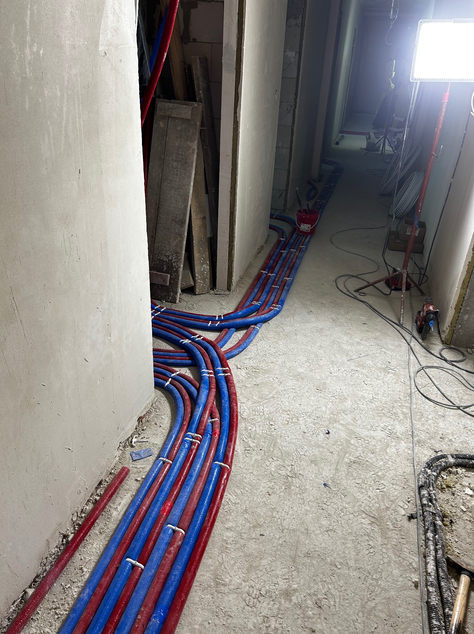 Instalacja rur grzewczych (czerwone i niebieskie) ułożonych wzdłuż ściany w surowym wnętrzu. Widoczne narzędzia i oświetlenie budowlane.