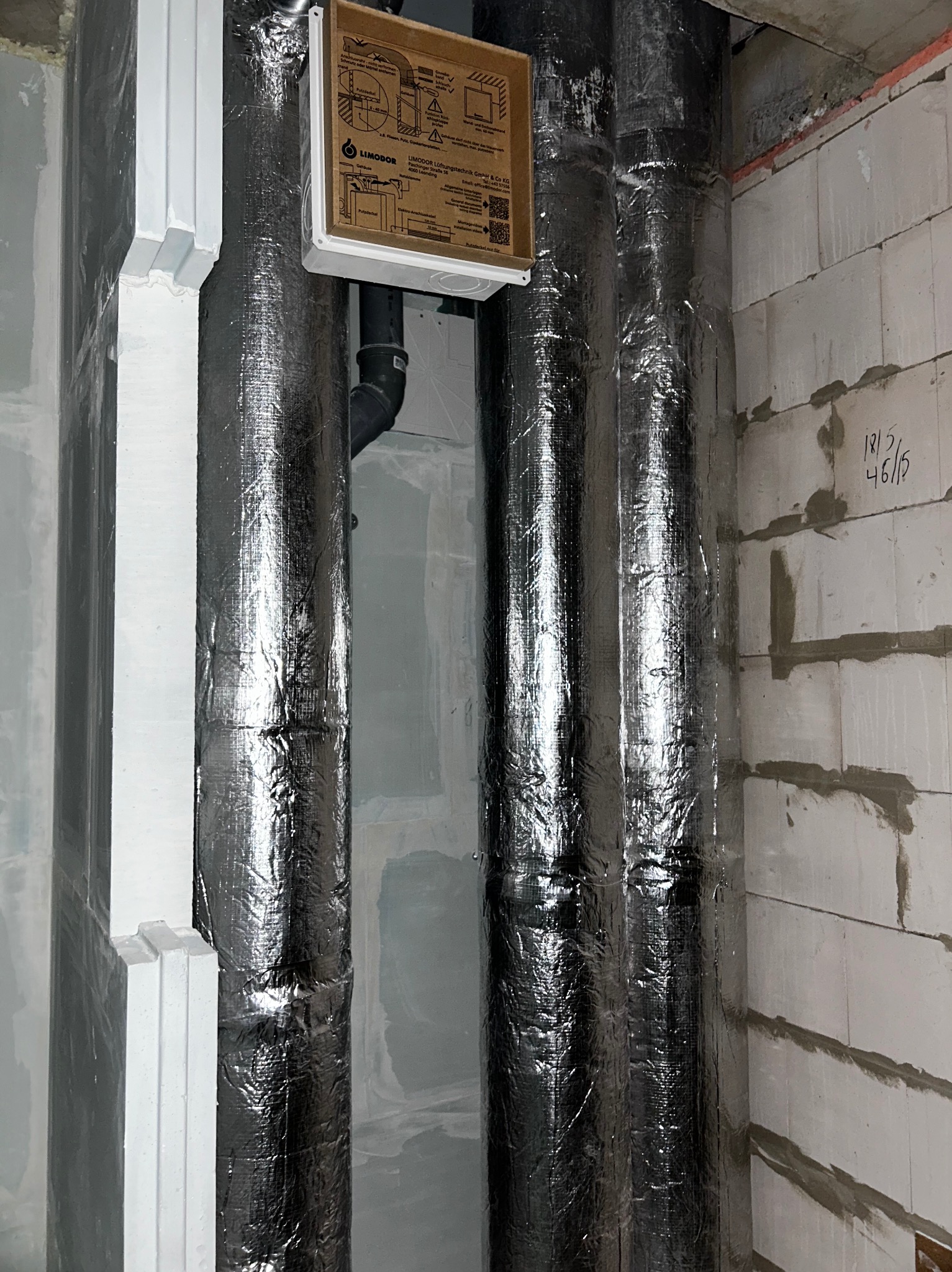 Dwie pionowe rury w srebrnej izolacji, z podłączonym przewodem i białą skrzynką Limodor na ścianie z bloczków. Instalacja w surowym stanie.
