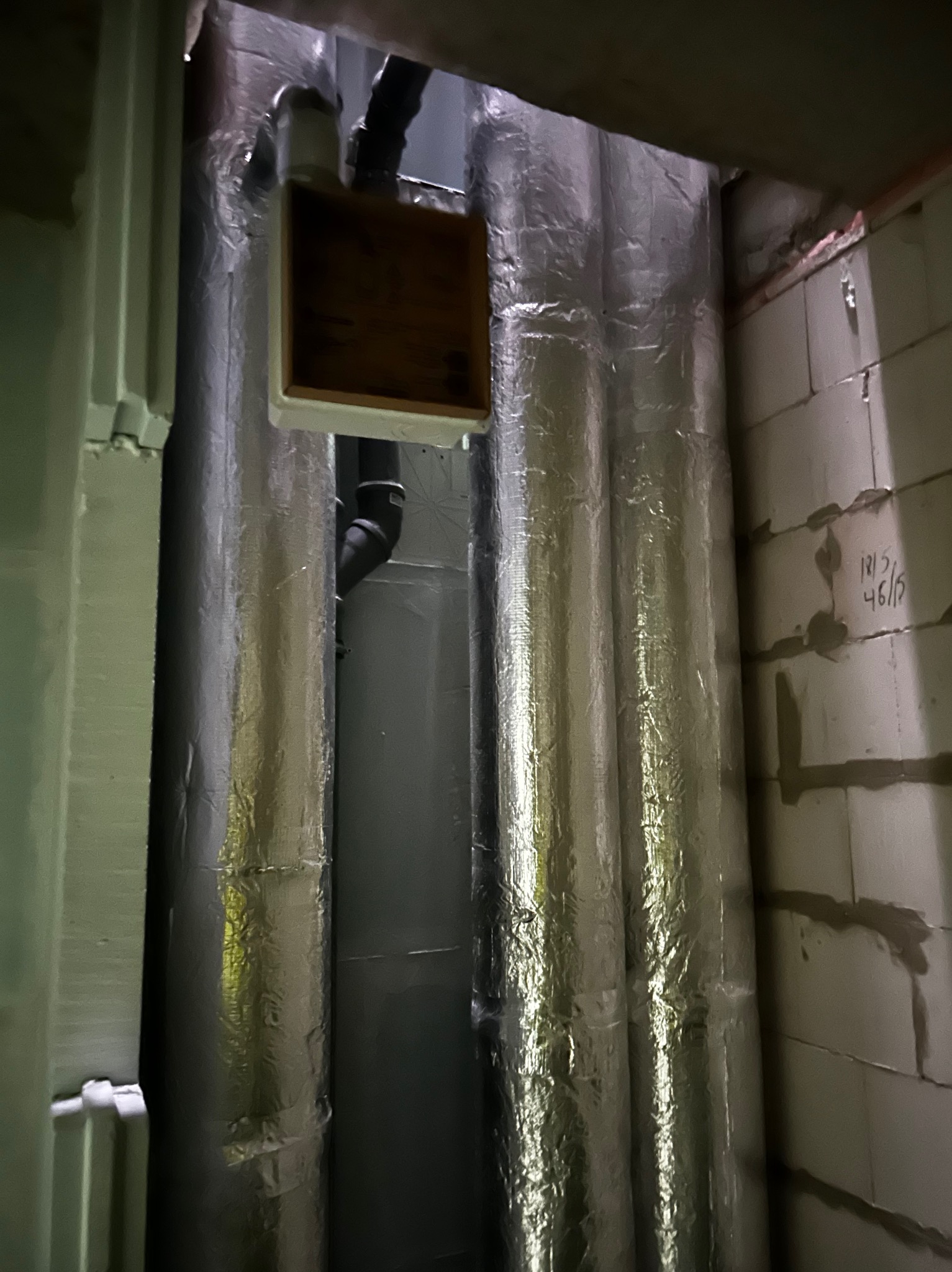 Izolowane rury wentylacyjne w piwnicy, owinięte folią aluminiową, z widocznymi elementami instalacji i fragmentem ściany z betonu komórkowego.