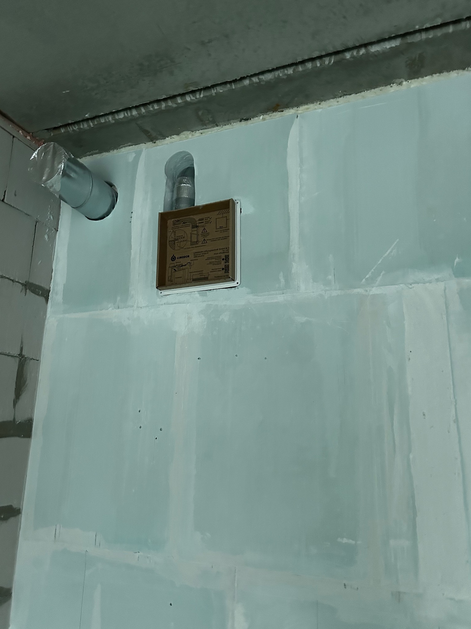 Szara ściana z płyt kartonowo-gipsowych z widocznymi elementami instalacji wentylacyjnej: rura z tworzywa sztucznego i otwór z kartonowym pudełkiem. Surowy stan wykończenia.