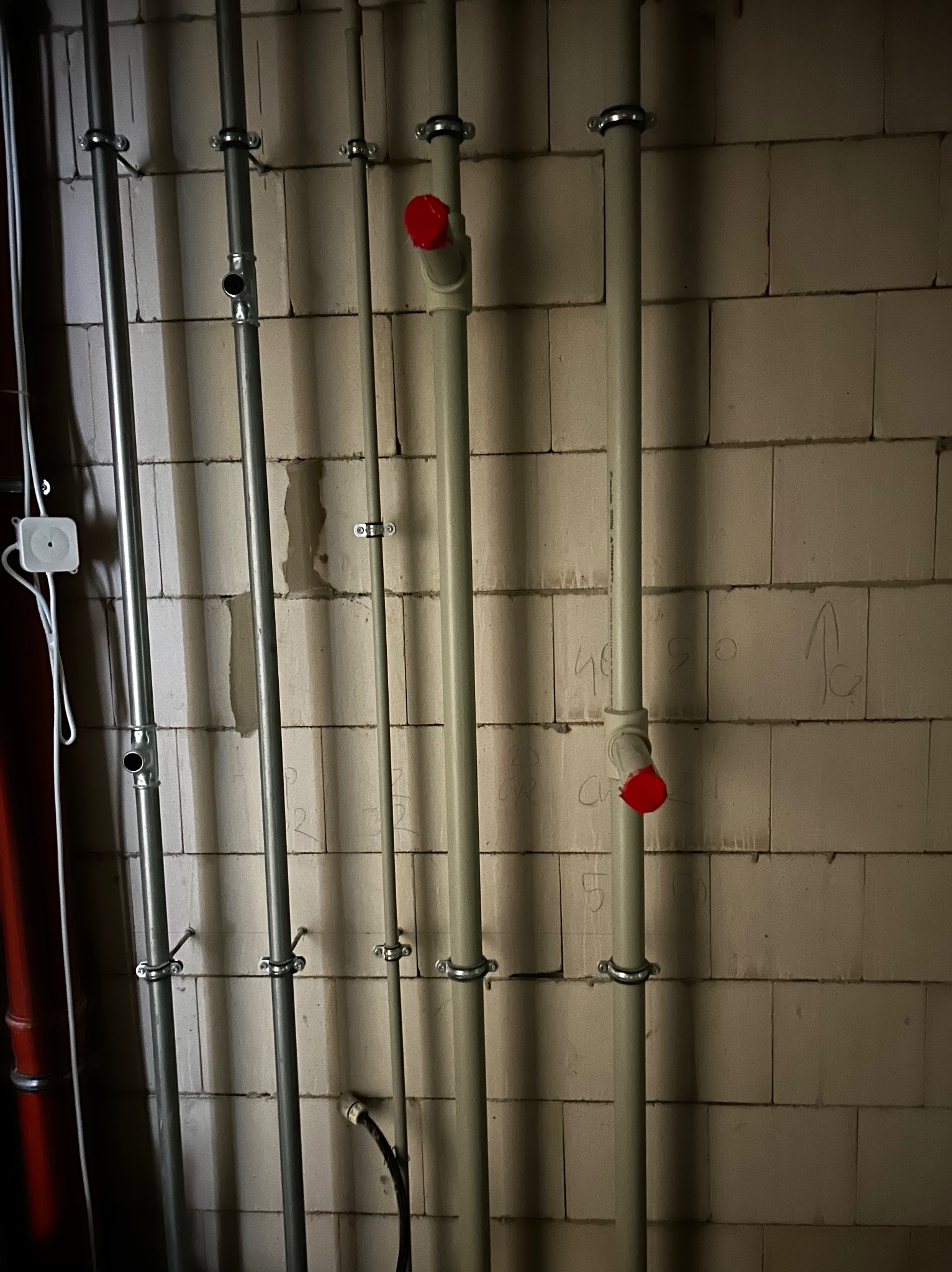Pionowe rury instalacyjne (metalowe i PCV z czerwonymi zaślepkami) przymocowane do ściany z betonu komórkowego. Widoczne uchwyty mocujące i fragment instalacji elektrycznej.