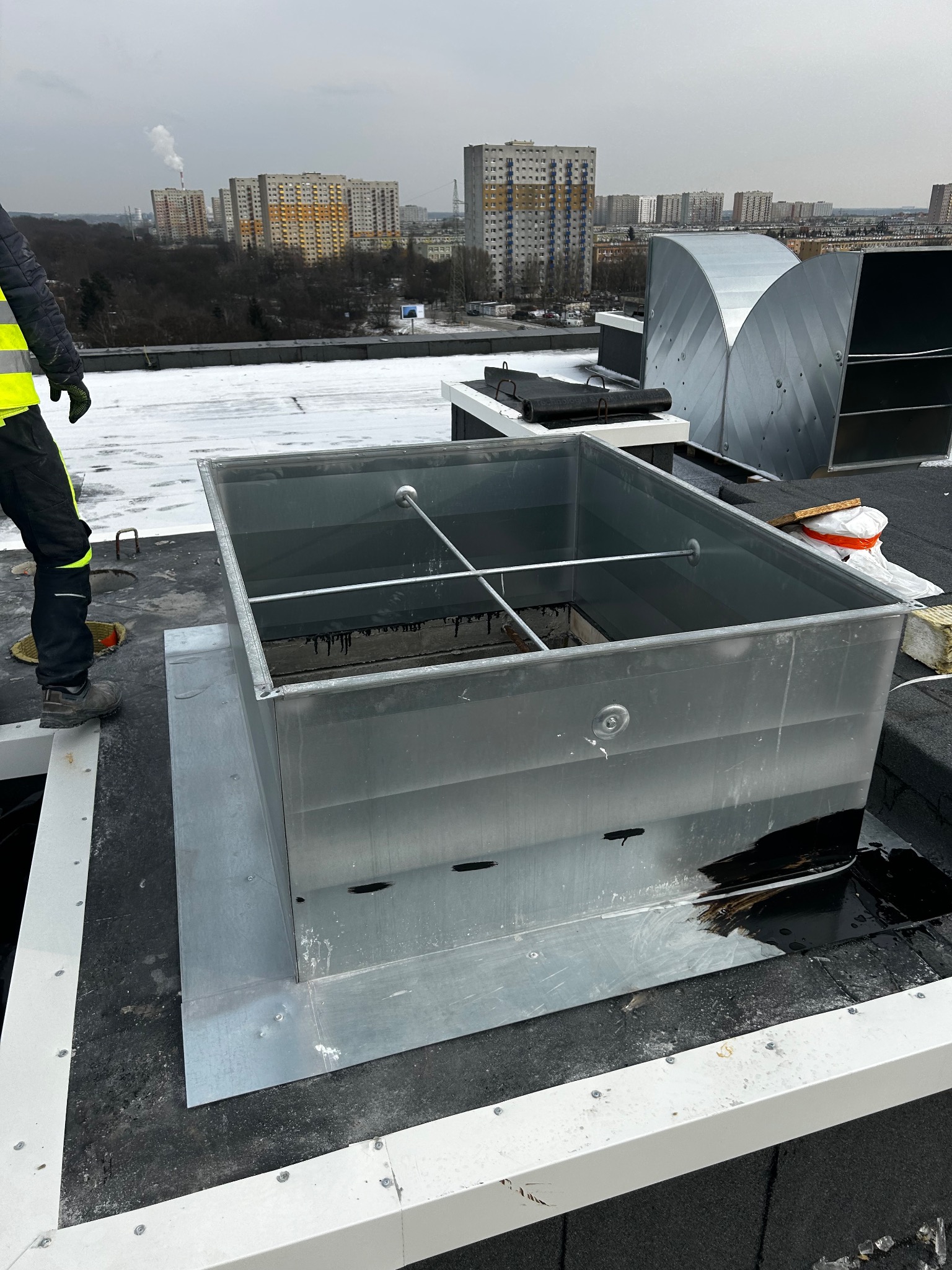 Montaż metalowej konstrukcji wentylacyjnej na dachu budynku. Widoczny pracownik w odblaskowej kamizelce. W tle zabudowa miejska i pochmurne niebo.