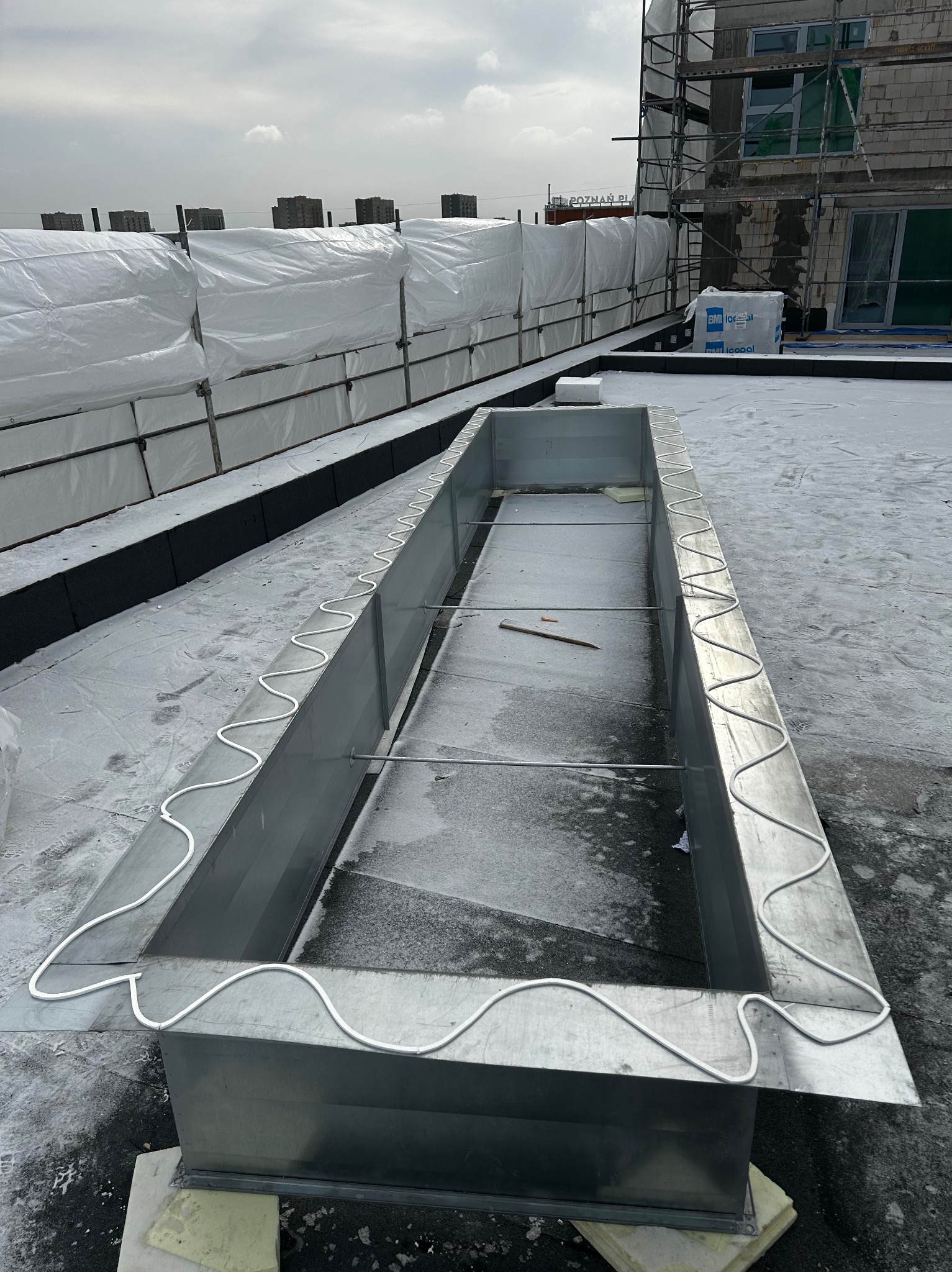 Metalowa obudowa na dachu budynku w trakcie montażu, zabezpieczona białym kablem grzewczym. W tle rusztowania i budynek w budowie, pochmurne niebo.