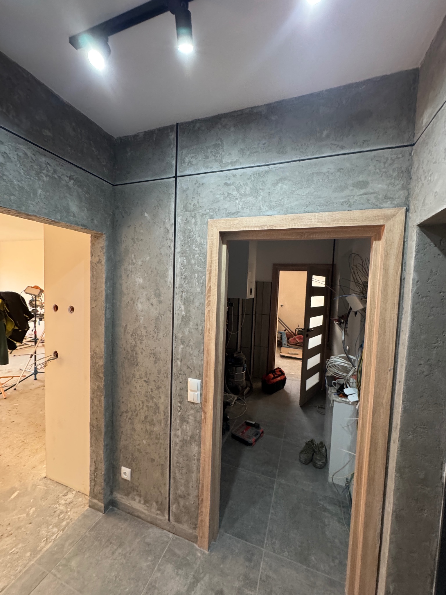 Korytarz w trakcie remontu: ściany wykończone szarą, betonową fakturą z czarnymi listwami, widoczne niedokończone instalacje elektryczne, ościeżnice drzwiowe z surowego drewna, podłoga wyłożona...