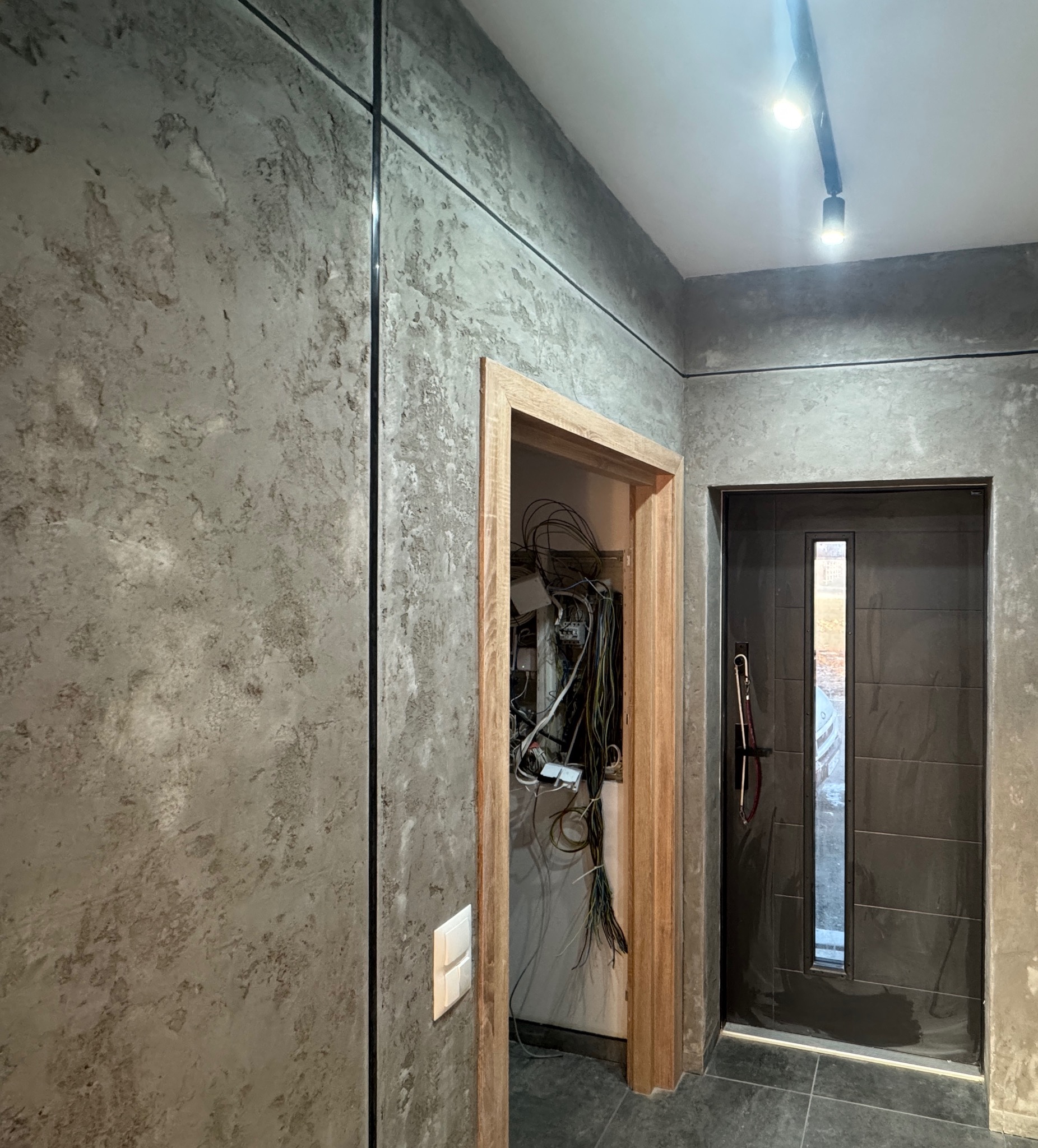 Korytarz w trakcie remontu, ściany z efektem betonu, widoczna instalacja elektryczna w otwartym przejściu i nowoczesne drzwi wejściowe.