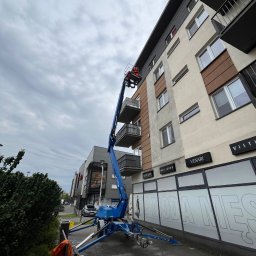 Usługi Wysokościowe - Dominik Lipski - Niebieski podnośnik koszowy przy budynku, osoba w koszu na wysokości balkonu. Widoczne okna i szyldy sklepów. Pochmurne niebo w tle.