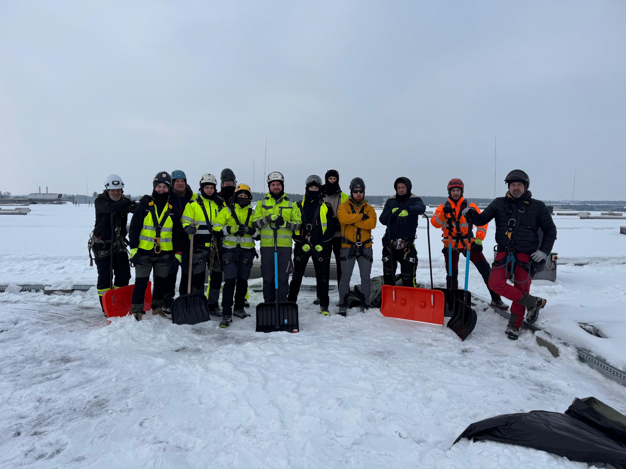 Grupa pracowników w kaskach i uprzężach, z łopatami na zasypanym śniegiem dachu, gotowa do pracy. W tle widoczny budynek i pochmurne niebo.