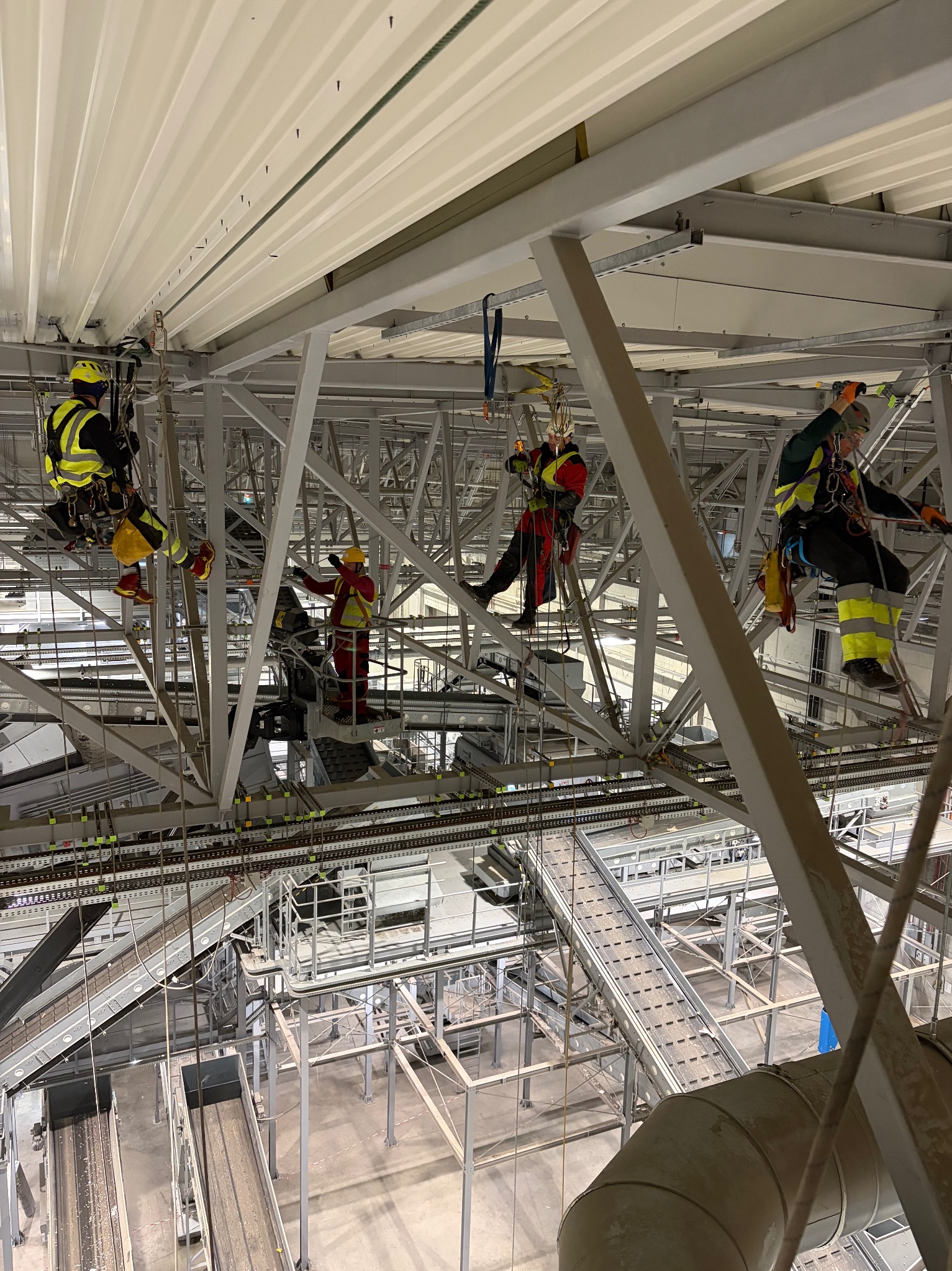 Pracownicy w uprzężach na konstrukcji hali, wykonujący zadania na wysokości. Widoczne elementy konstrukcyjne i systemy transportowe. Ujęcie z dołu.