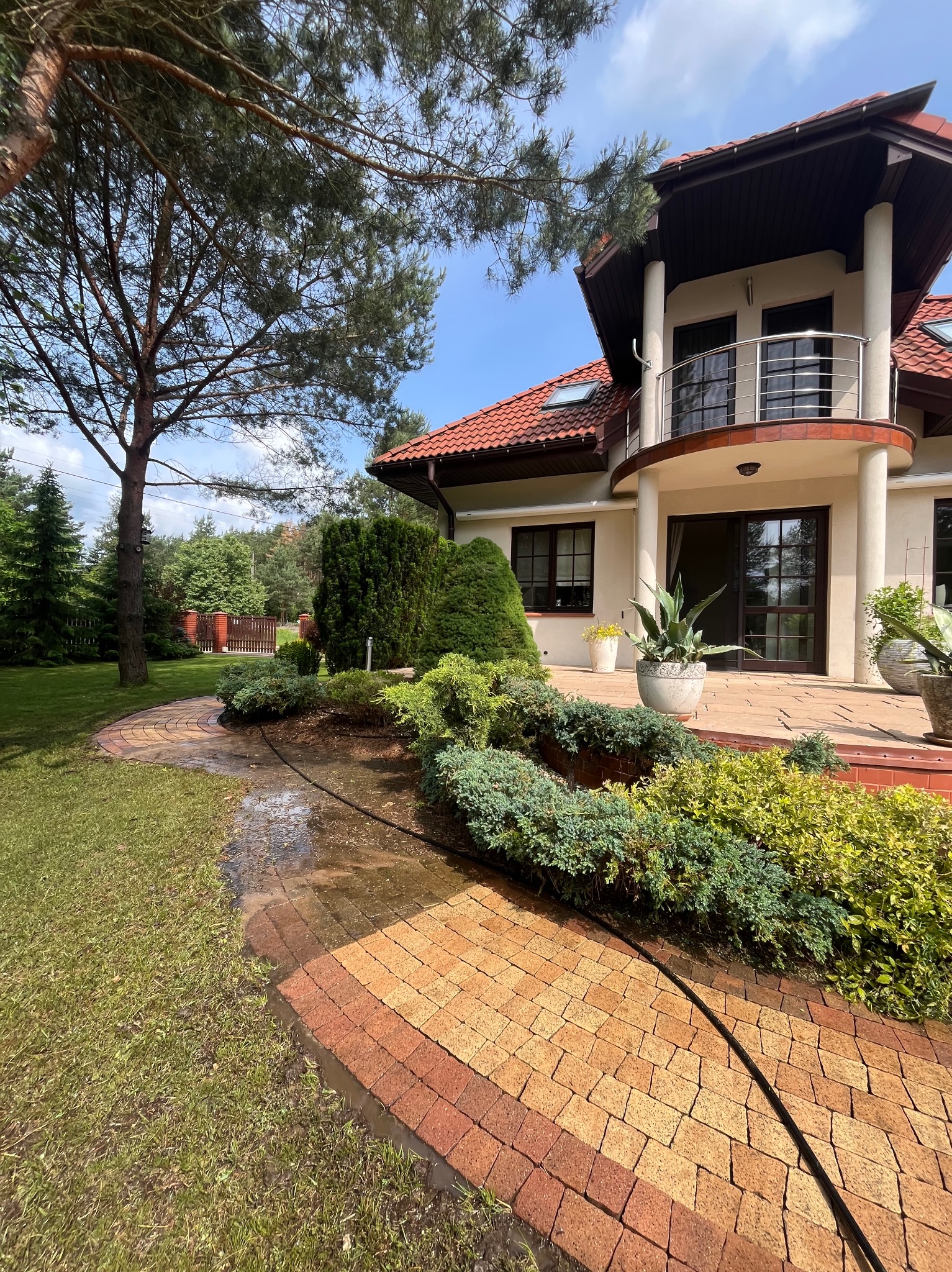 Mycie kostki brukowej na posesji domu jednorodzinnego. Wąż ciśnieniowy leży na mokrej kostce. Widoczne krzewy, trawnik i fragment budynku z balkonem.