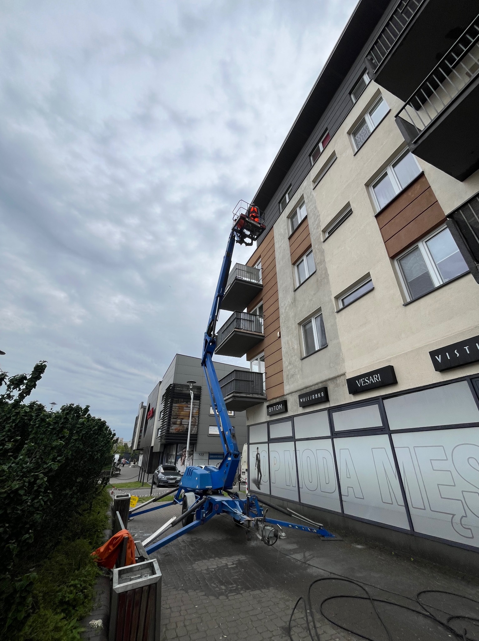 Niebieski podnośnik koszowy przy budynku, osoba w koszu na wysokości balkonu. Widoczne okna i szyldy sklepów. Pochmurne niebo w tle.