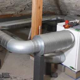 Montaż rekuperatora WANAS w surowym wnętrzu poddasza z izolacją natryskową, widoczne metalowe rury wentylacyjne, narzędzia i akcesoria na obudowie urządzenia.