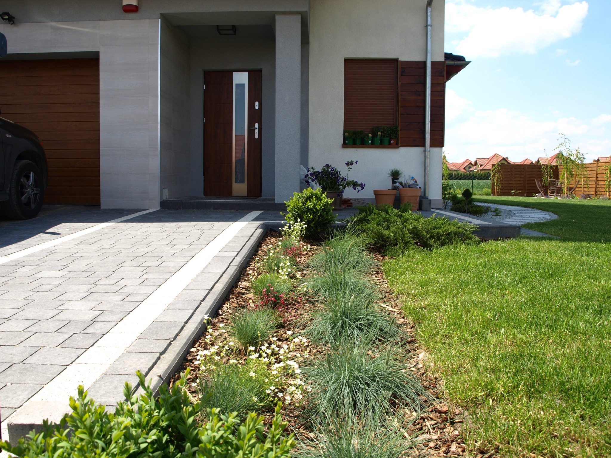 Ogród przed domem z rabatą kwiatową wzdłuż ścieżki z kostki brukowej, trawnik, drewniane ogrodzenie w tle.