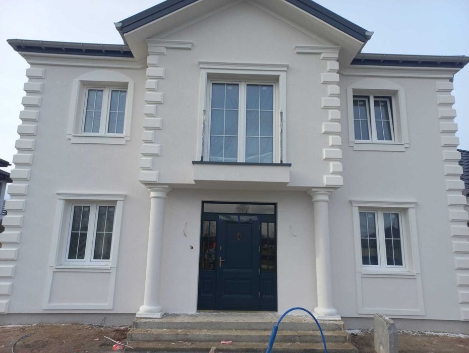 Wykończona elewacja dwupiętrowego domu z białym tynkiem, dekoracyjnymi kolumnami i obramowaniami okien, widoczne niebieskie rury na schodach wejściowych.