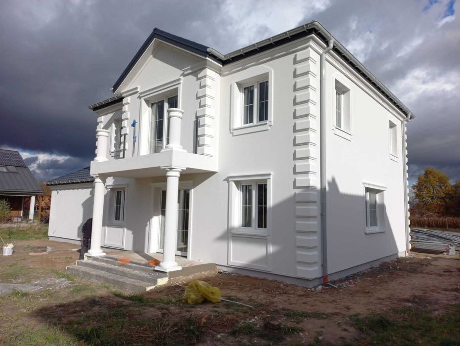Biały, dwukondygnacyjny dom z balkonem wspartym na kolumnach, wykończony sztukaterią, na tle pochmurnego nieba, w trakcie prac wykończeniowych elewacji.