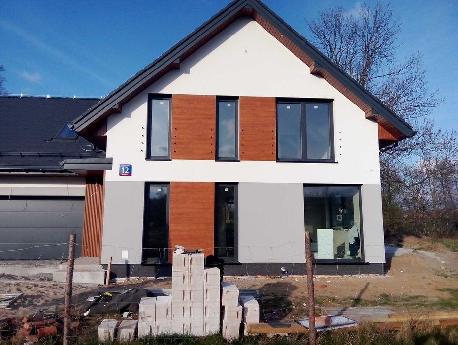 Nowoczesny dom jednorodzinny w trakcie wykańczania elewacji, z fragmentami drewnianej okładziny, szarą dolną częścią i białym wykończeniem górnej części, widoczne materiały budowlane przed budynkiem.