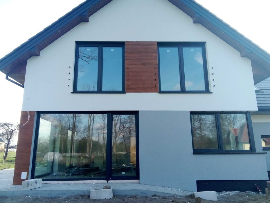 Dwukondygnacyjny dom z jasną elewacją, drewnianymi akcentami i dużymi oknami z czarnymi ramami, widok z zewnątrz na etapie wykończenia.