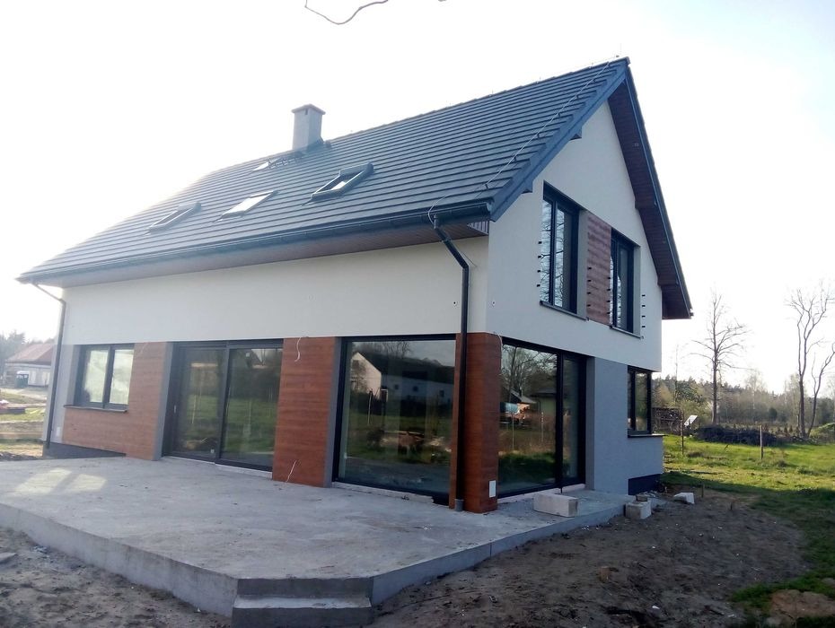 Nowoczesny dom jednorodzinny z jasną elewacją, ciemną dachówką i dużymi oknami przesuwnymi, widoczne fragmenty elewacji wykończone imitacją drewna, surowy betonowy taras przed budynkiem.