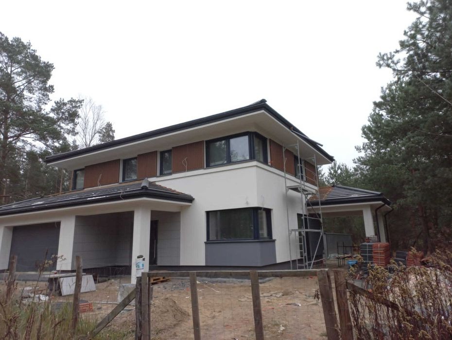 Dwukondygnacyjny dom z białymi ścianami i brązowymi panelami na górnej kondygnacji, widoczne rusztowanie przy elewacji, teren wokół domu częściowo uporządkowany, w tle las.