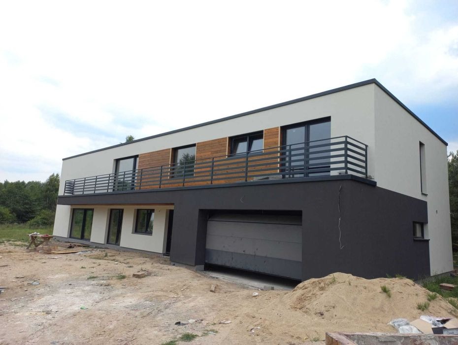 Dwukondygnacyjny dom z płaskim dachem, wykończony jasnoszarym tynkiem na górnej kondygnacji i ciemnoszarym na dolnej, z drewnianymi panelami na balkonie i metalowymi balustradami, widoczny plac...