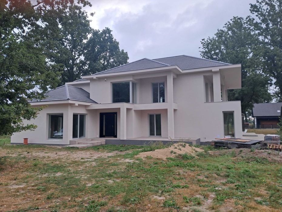 Dwukondygnacyjny dom jednorodzinny z płaskim dachem, elewacja w jasnym kolorze, duże okna, widoczne ślady prac wykończeniowych wokół budynku, na pierwszym planie trawa i drzewa.