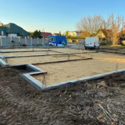 Przygotowany teren pod fundamenty z widocznymi zbrojeniami, piaskiem i izolacją, na tle zabudowy jednorodzinnej i zaparkowanego białego samochodu dostawczego.