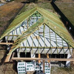 AK INVEST Anna Karaś - Wysoki kąt widzenia na budowę domu w stanie surowym otwartym z widoczną konstrukcją dachu z impregnowanego drewna, palety z bloczkami H+H przed budynkiem i dwoma osobami w tle wzdłuż ogrodzenia.