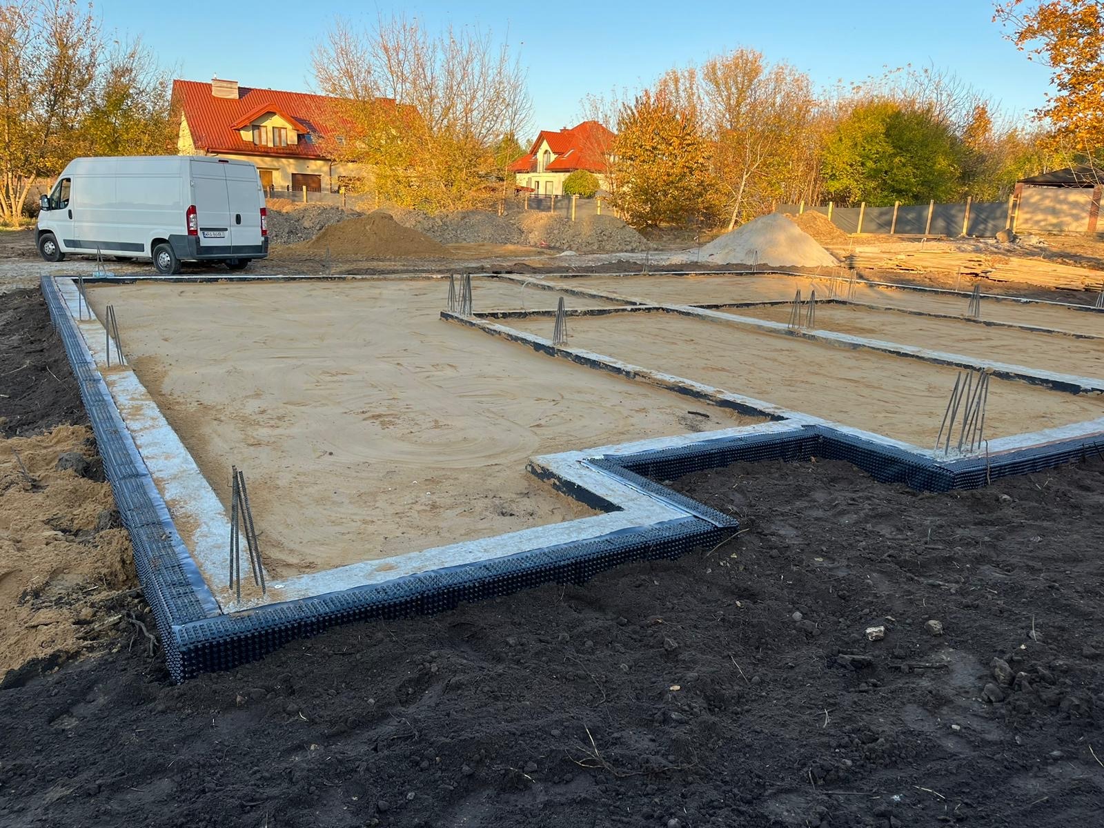 Przygotowany teren pod budowę domu jednorodzinnego z widocznymi ławami fundamentowymi zabezpieczonymi folią kubełkową i wystającymi prętami zbrojeniowymi, w tle zaparkowany biały bus i dwa domy...