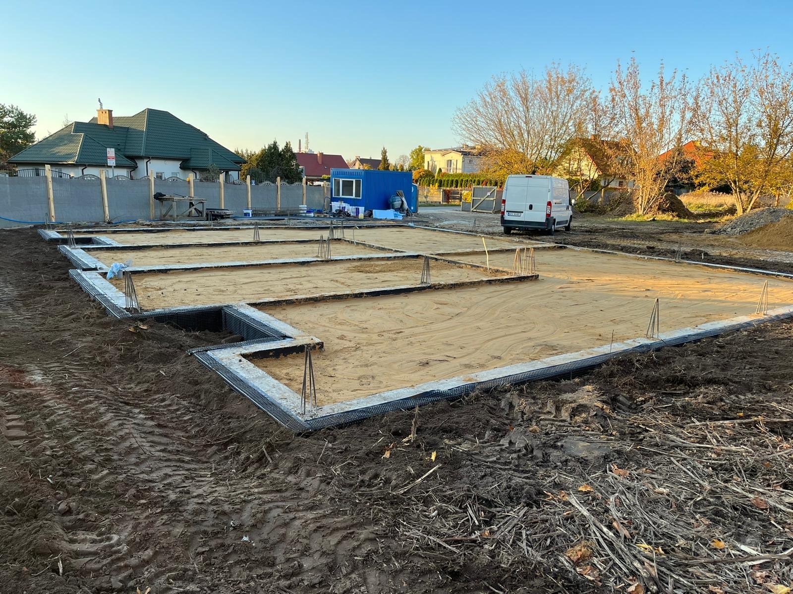 Przygotowany teren pod fundamenty z widocznymi zbrojeniami, piaskiem i izolacją, na tle zabudowy jednorodzinnej i zaparkowanego białego samochodu dostawczego.