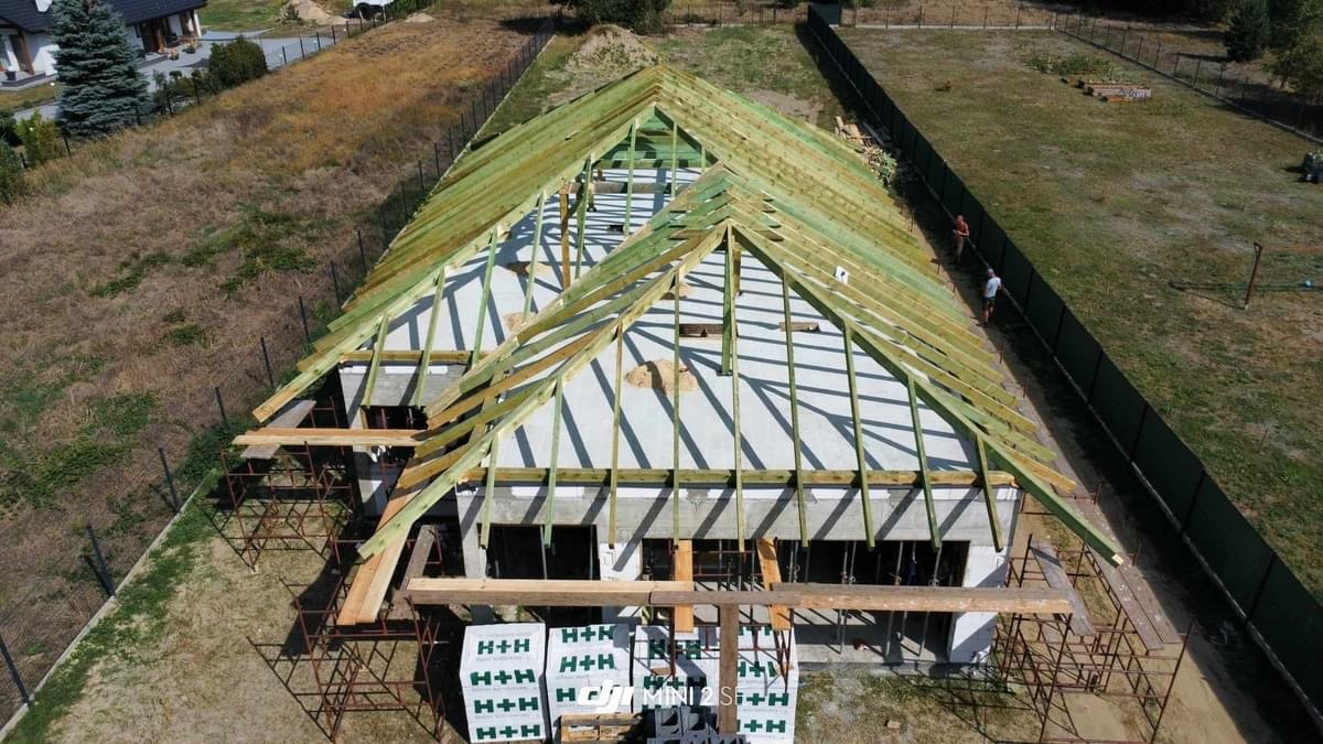 Wysoki kąt widzenia na budowę domu w stanie surowym otwartym z widoczną konstrukcją dachu z impregnowanego drewna, palety z bloczkami H+H przed budynkiem i dwoma osobami w tle wzdłuż ogrodzenia.