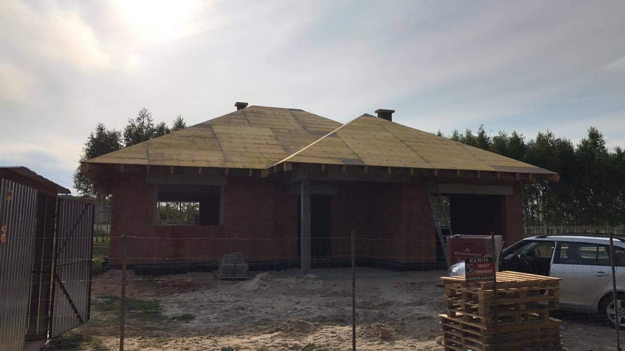 Niedokończony budynek jednorodzinny z brązowych pustaków, z prowizorycznym dachem z desek, widoczna drabina oparta o ścianę garażu, teren budowy z paletami i zaparkowanym samochodem osobowym.