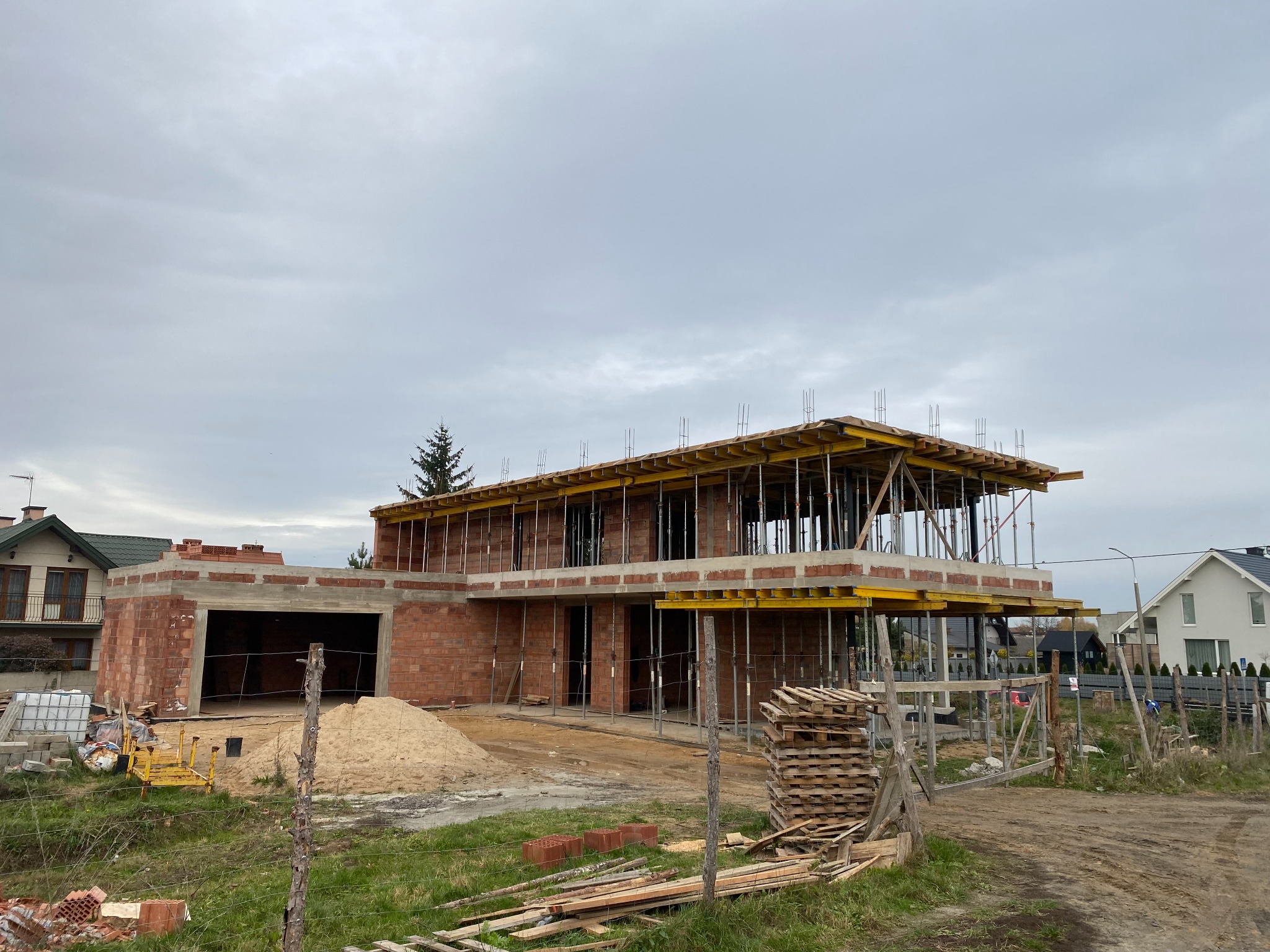 Dwukondygnacyjny dom w trakcie budowy, z widocznymi elementami szalunków, ścianami z czerwonej cegły i stertą piasku przed garażem, na tle pochmurnego nieba.