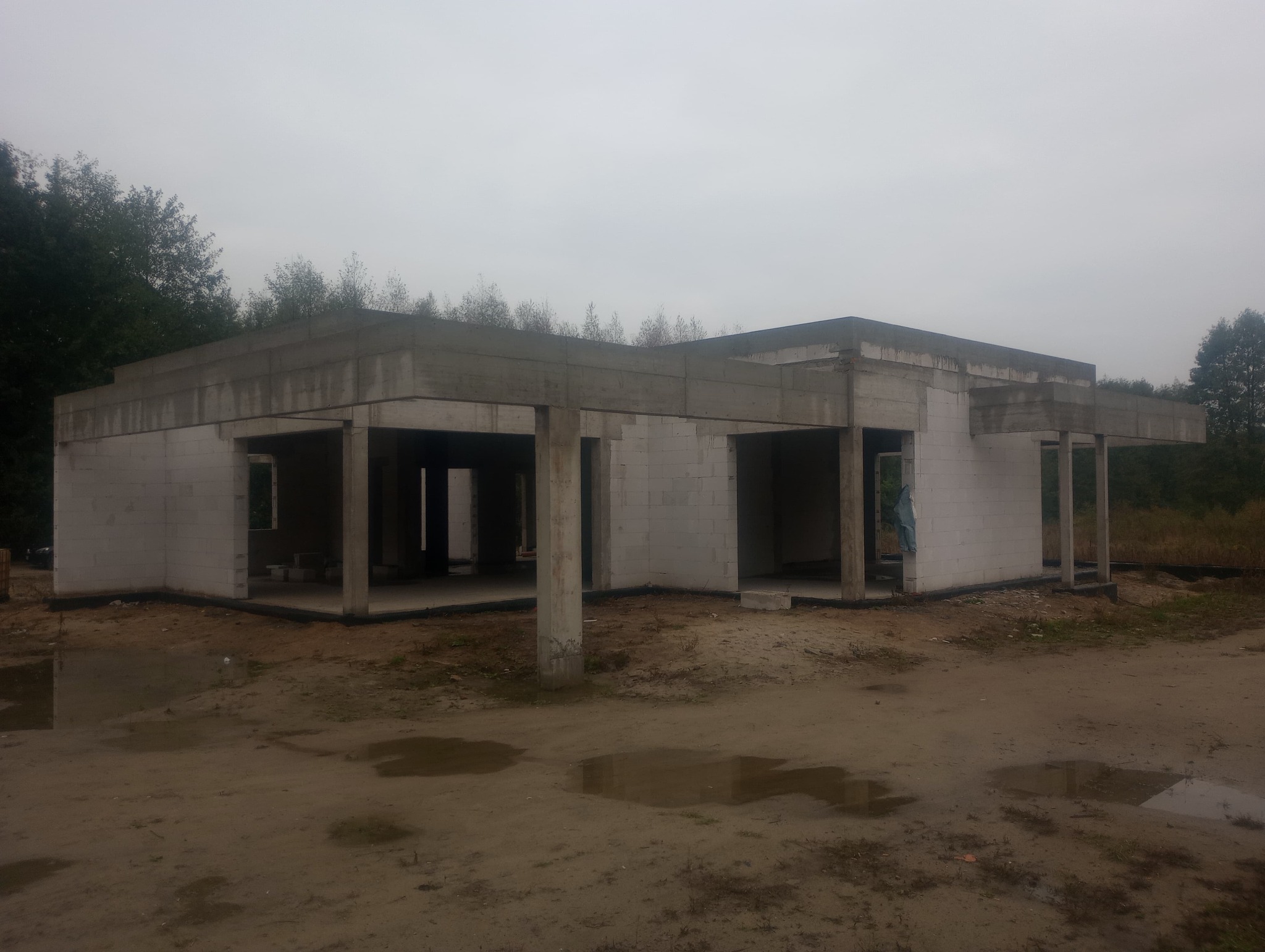 Surowy stan parterowego budynku z betonu komórkowego pod szarym niebem, widoczne kolumny i nadproża, teren wokół z błotnistymi kałużami.