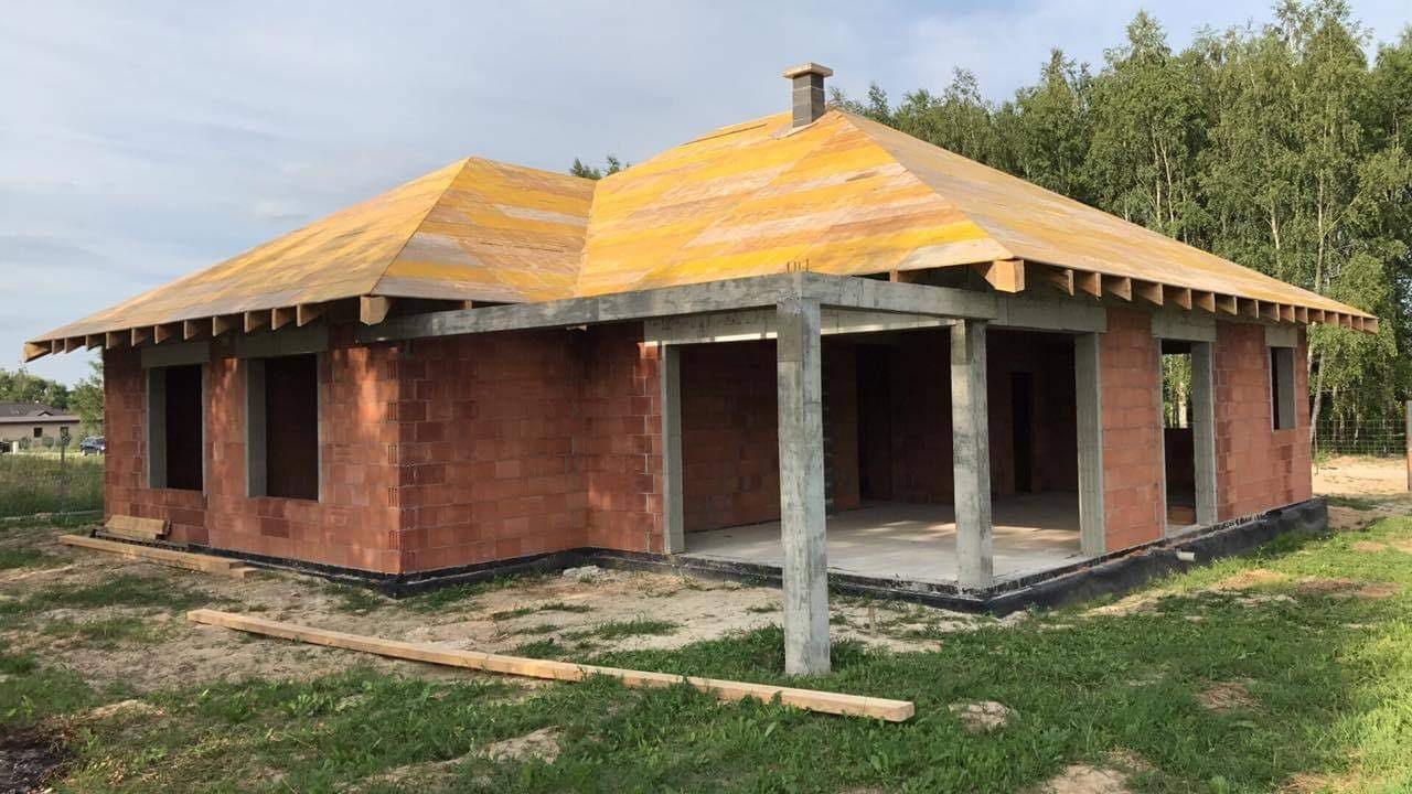 Surowy stan domu murowanego z keramzytu z żółtą membraną dachową, widoczne betonowe słupy werandy i fragment fundamentu z izolacją przeciwwilgociową, w tle las.