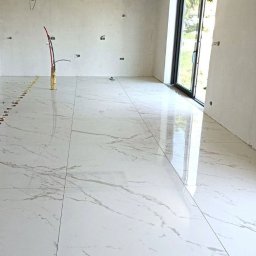 Katarzyna Dublasiewicz - Układanie białych płytek imitujących marmur na podłodze w jasnym, przestronnym pomieszczeniu z dużym oknem balkonowym; widoczne kable wystające ze ściany i listwy dystansowe pomiędzy płytkami.