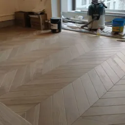 Ułożona w jodełkę jasna podłoga drewniana, widoczne narzędzia i materiały budowlane przy oknie, w tle budynek.