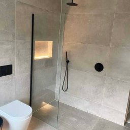 Katarzyna Dublasiewicz - Nowoczesna łazienka z szarymi płytkami, kabiną prysznicową typu walk-in z czarnymi elementami armatury, podświetlaną wnęką w ścianie i podwieszaną toaletą.