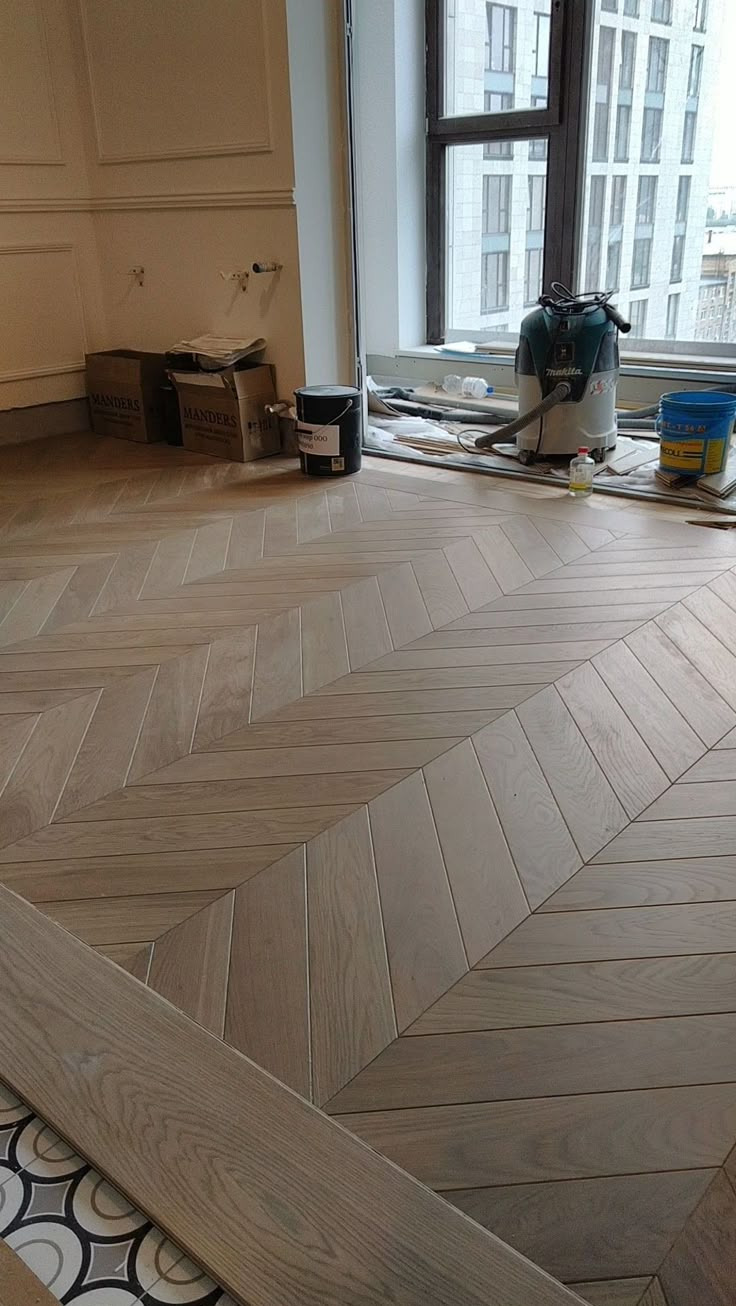 Ułożona w jodełkę jasna podłoga drewniana, widoczne narzędzia i materiały budowlane przy oknie, w tle budynek.