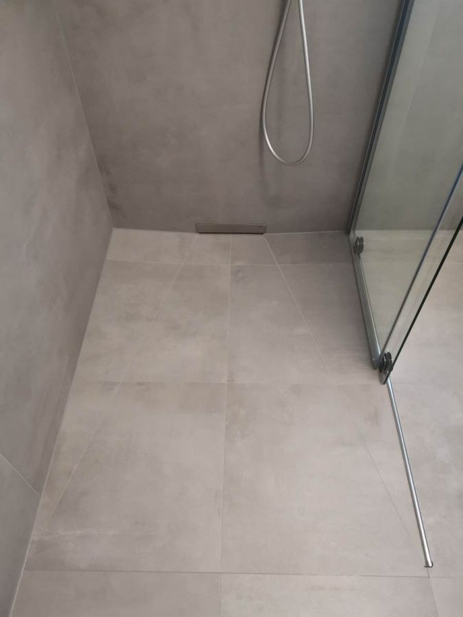 Nowoczesny prysznic walk-in z odpływem liniowym, wykończony szarą, matową glazurą wielkoformatową i szklaną kabiną z chromowanymi okuciami.