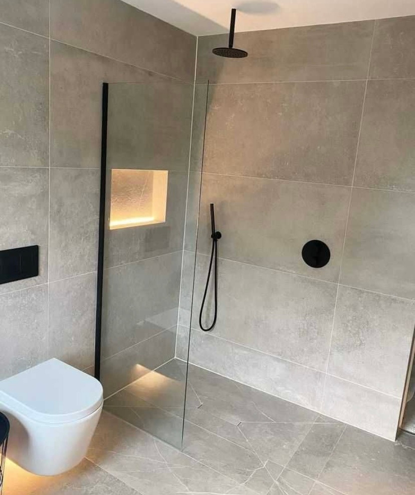 Nowoczesna łazienka z szarymi płytkami, kabiną prysznicową typu walk-in z czarnymi elementami armatury, podświetlaną wnęką w ścianie i podwieszaną toaletą.