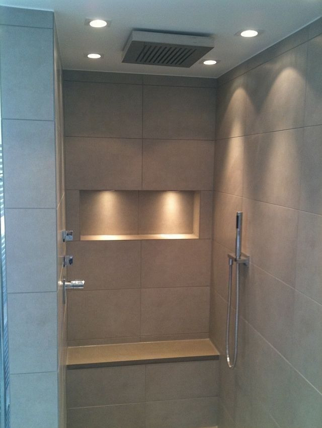 Nowoczesna kabina prysznicowa z szarymi płytkami, wbudowaną półką podświetloną punktowo, deszczownicą sufitową i siedziskiem z tego samego materiału co ściany.