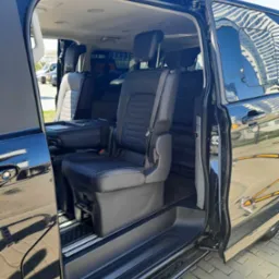 Wnętrze czarnego, luksusowego vana z otwartymi drzwiami przesuwnymi, ukazujące skórzane fotele z zagłówkami i pasami bezpieczeństwa, z widocznym budynkiem odbijającym się w lakierze.