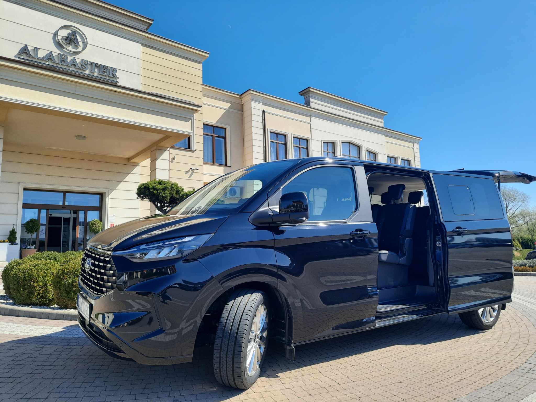 Czarny van z otwartymi drzwiami pasażera i bagażnika zaparkowany przed budynkiem z napisem 'ALABASTER'.