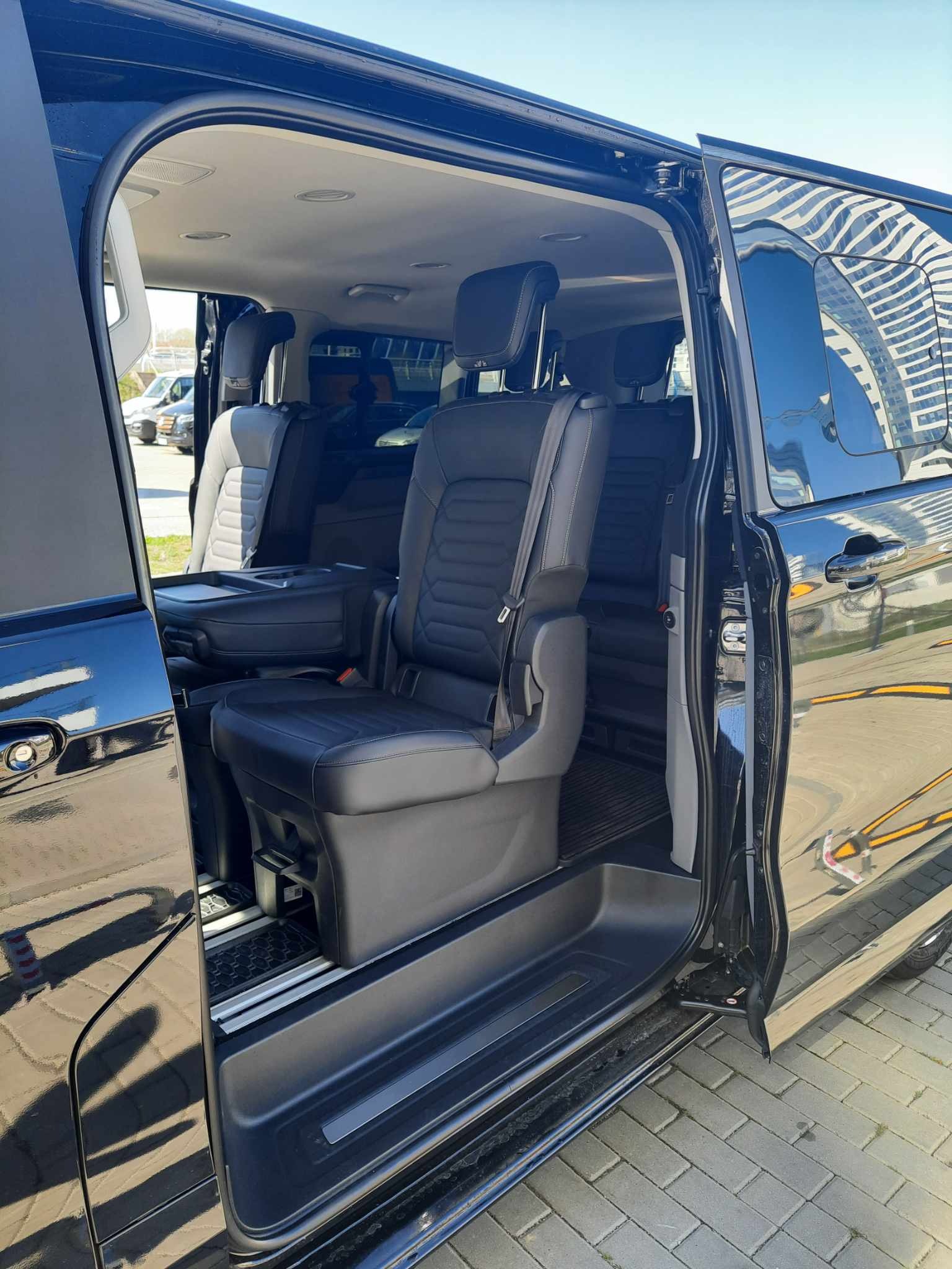 Wnętrze czarnego, luksusowego vana z otwartymi drzwiami przesuwnymi, ukazujące skórzane fotele z zagłówkami i pasami bezpieczeństwa, z widocznym budynkiem odbijającym się w lakierze.